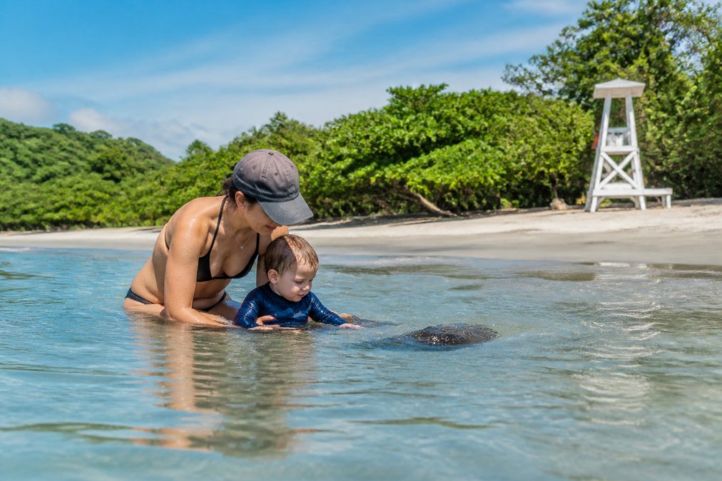 Parent supervising a toddler in shallow beach water near shore, with lifeguard tower visible in distance and calm shoreline conditions
