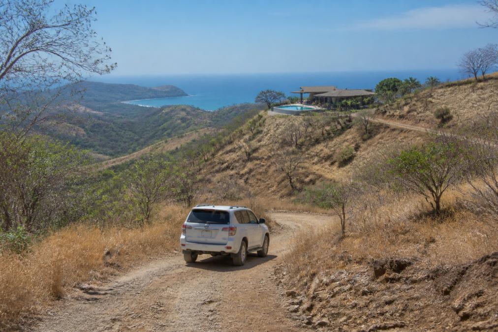 A white 4x4 SUV driving on a rugged dirt road through dry, golden Guanacaste hills toward a glimpse of the Pacific coast, a modern luxury villa with a pool visible on a ridge in the distance