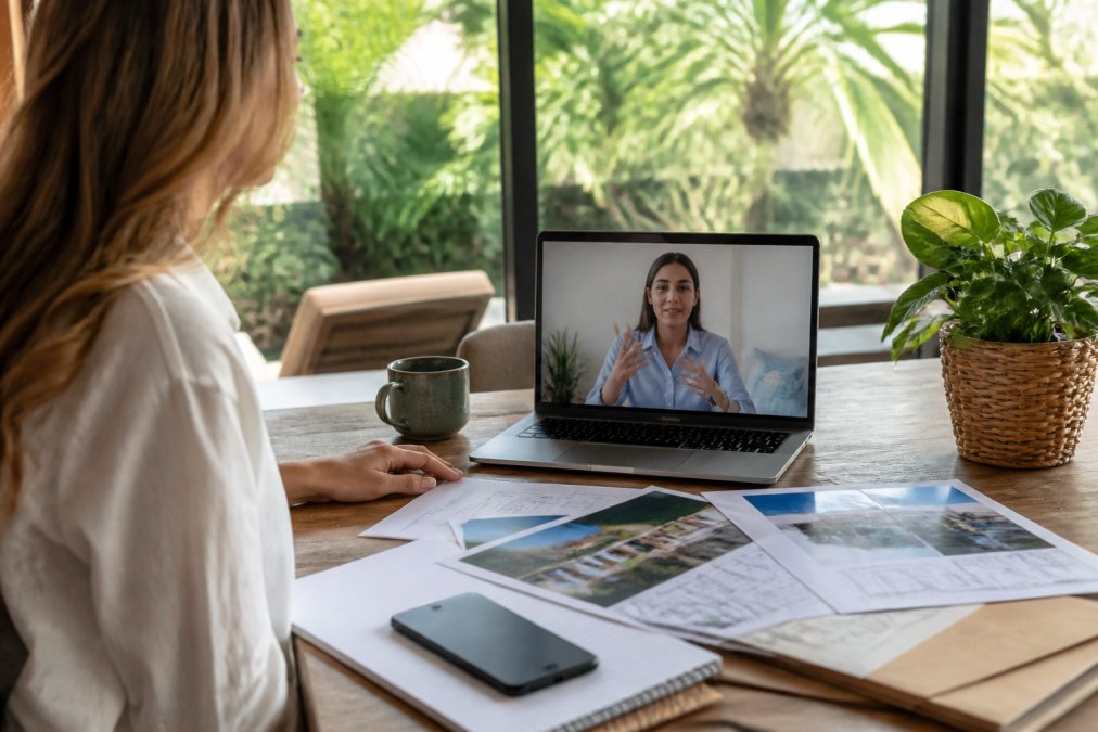 Remote property owner on a video call with a property manager, reviewing documents and discussing property management details on a laptop in a home office setting, natural lighting from windows