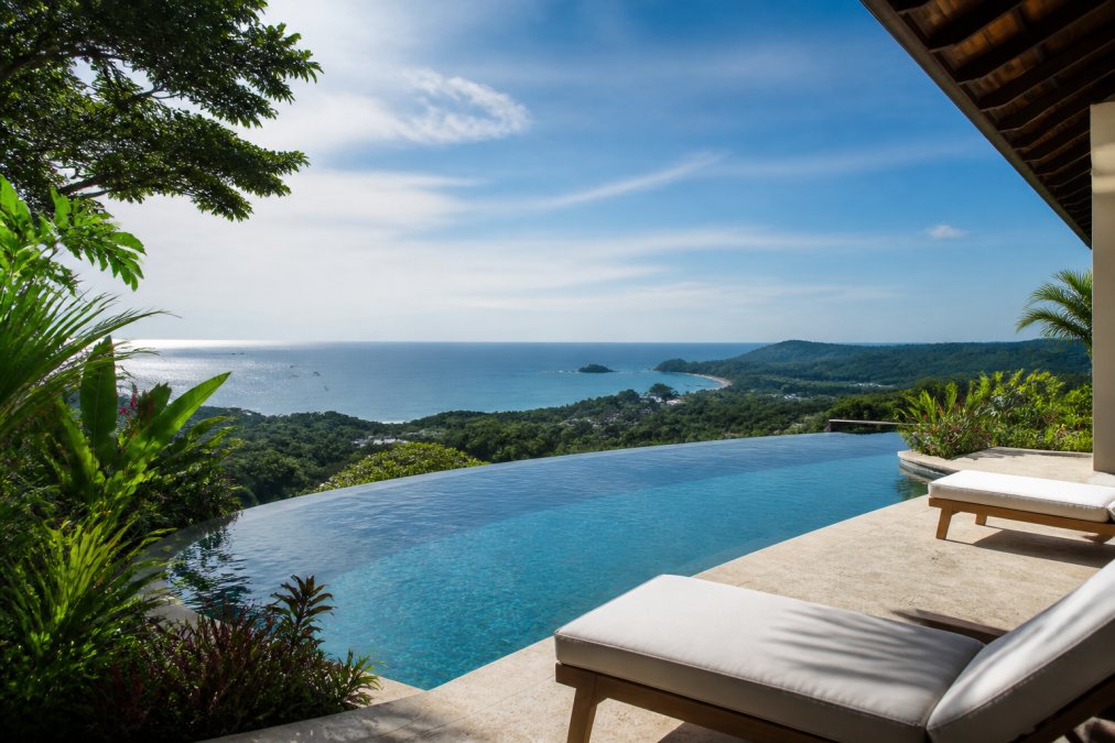 An infinity-edge pool at a luxury villa overlooking the Pacific Ocean in Guanacaste, with lounge chairs and tropical landscaped gardens in the foreground